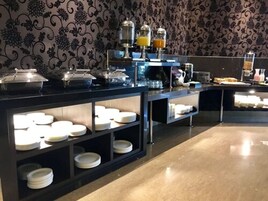Una mesa de buffet con diversos alimentos y bebidas, incluyendo una máquina de café y recipientes de vidrio con zumo de naranja.
