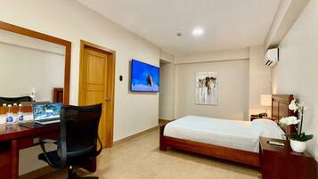 A hotel room with a bed, a desk with a laptop, a chair, a TV, and a painting on the wall.