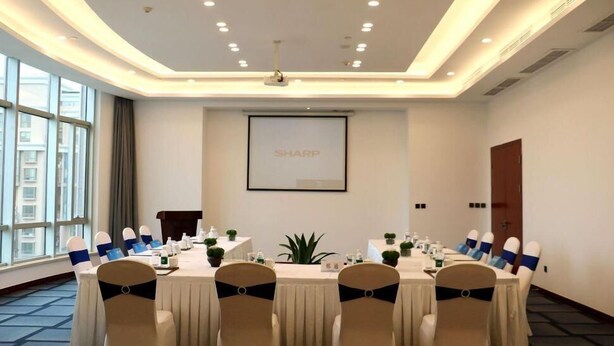 A conference room with a large table, chairs with blue sashes, a projector screen with 'SHARP' on it, and a window with a view of buildings.