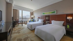 A hotel room with two beds, a desk, a chair, a TV, and a large window offering a city view.