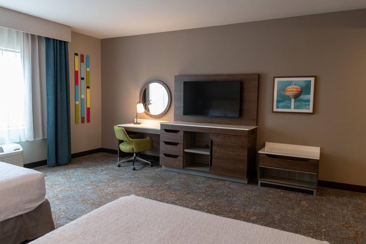Habitación de hotel con cama, escritorio con televisor, silla, espejo y un cuadro en la pared.