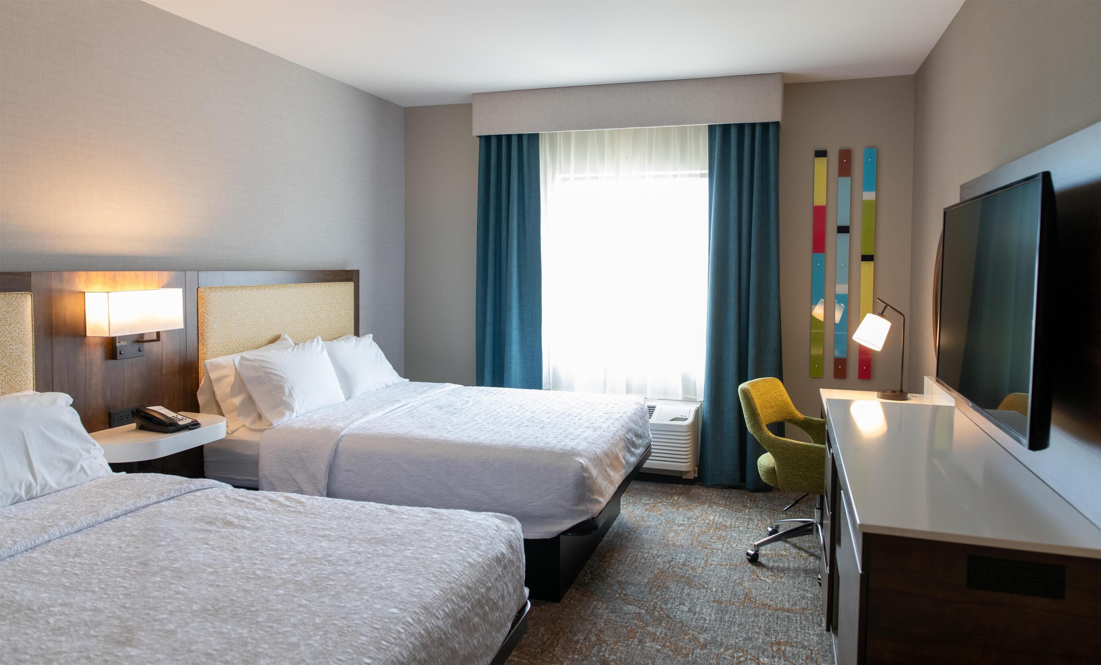 A hotel room with two beds, a desk with a TV, a chair, and a window with curtains.