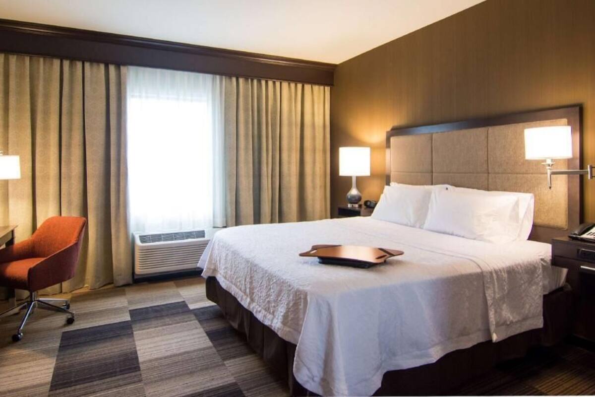 Habitación de hotel con cama, escritorio, una silla naranja, dos lámparas de noche y una ventana con cortinas.