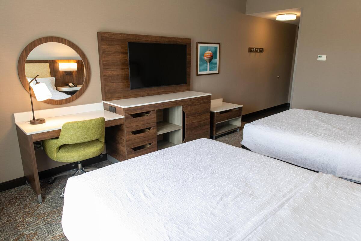 Habitación de hotel con escritorio, televisión, una silla verde, dos camas y un cuadro en la pared.