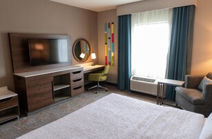 Habitación de hotel con cama, escritorio con silla, televisor, espejo, lámpara, ventana con cortinas y aire acondicionado instalado en la pared.