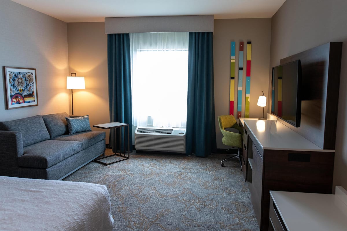 Habitación de hotel con sofá, escritorio, televisión de pantalla plana, lámpara y ventana grande con cortinas.