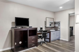 Una habitación de hotel moderna con un escritorio de madera oscura, un televisor de pantalla plana, una silla, una pequeña kitchenette y un espejo grande.