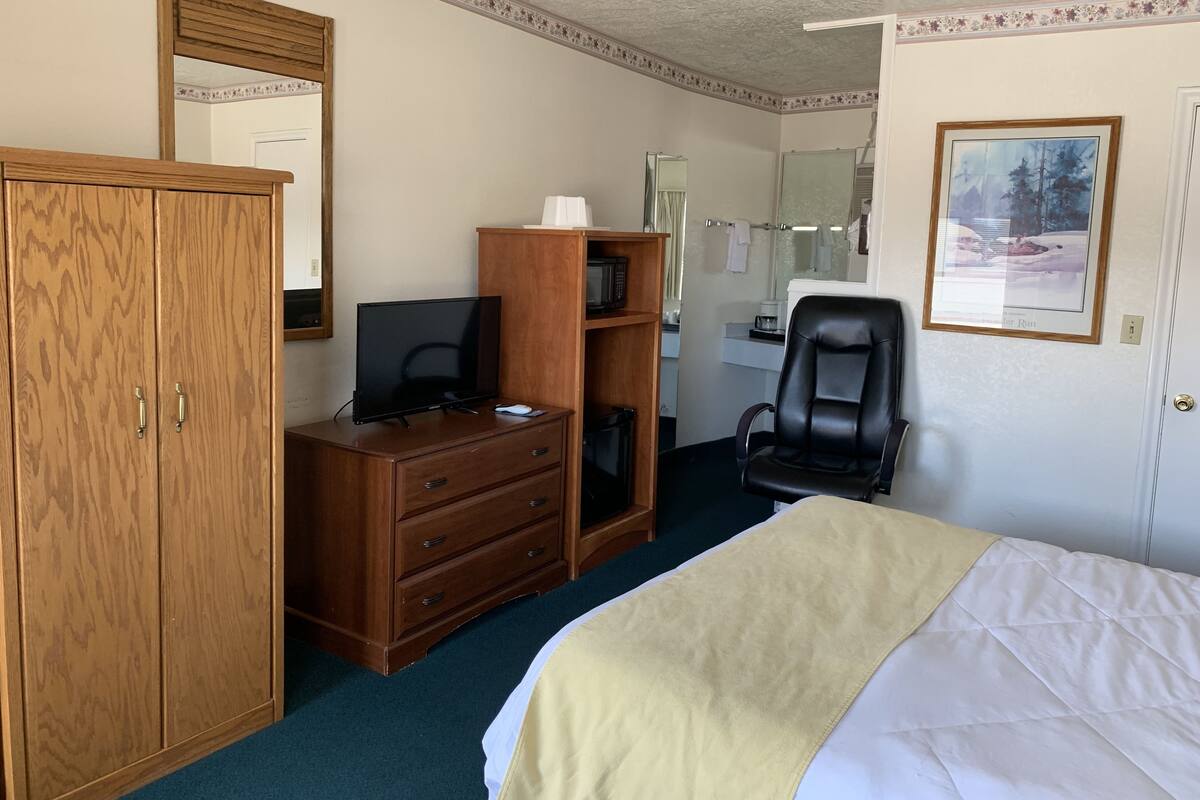 A hotel room with a bed, a wooden wardrobe, a desk with a computer, a chair, and a mirror.