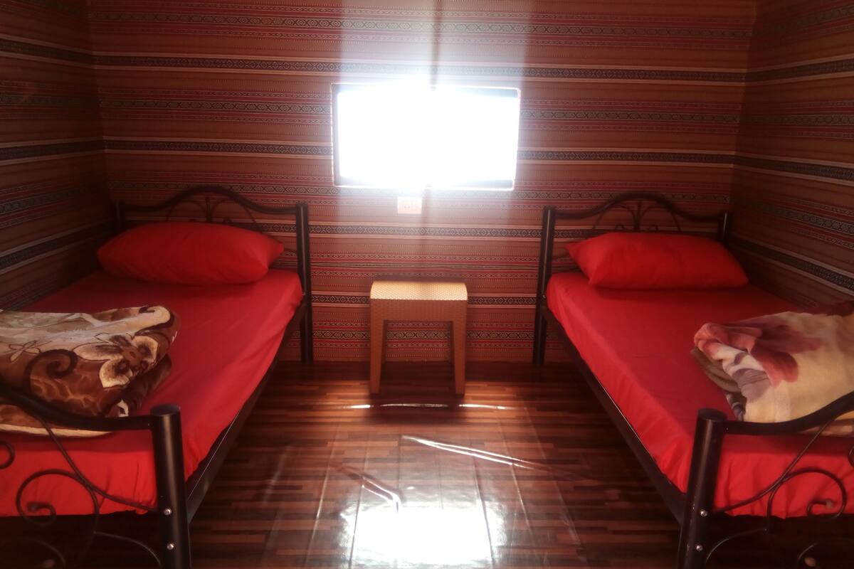 A small room with two beds, each with a red mattress and a brown headboard. There is a small table in the center with a light-colored top.