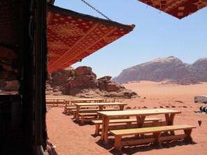 A desert landscape with rocky formations and a clear sky. There are wooden picnic tables arranged in the foreground. - Wadi Rum Caravan Camp (Wadi Rum)