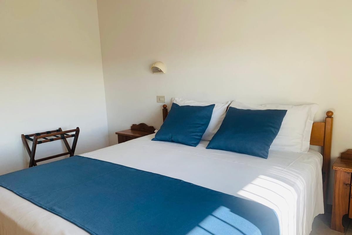 A neatly made bed with a blue bedspread and pillows, a wooden nightstand with a lamp, and a chair against the wall.