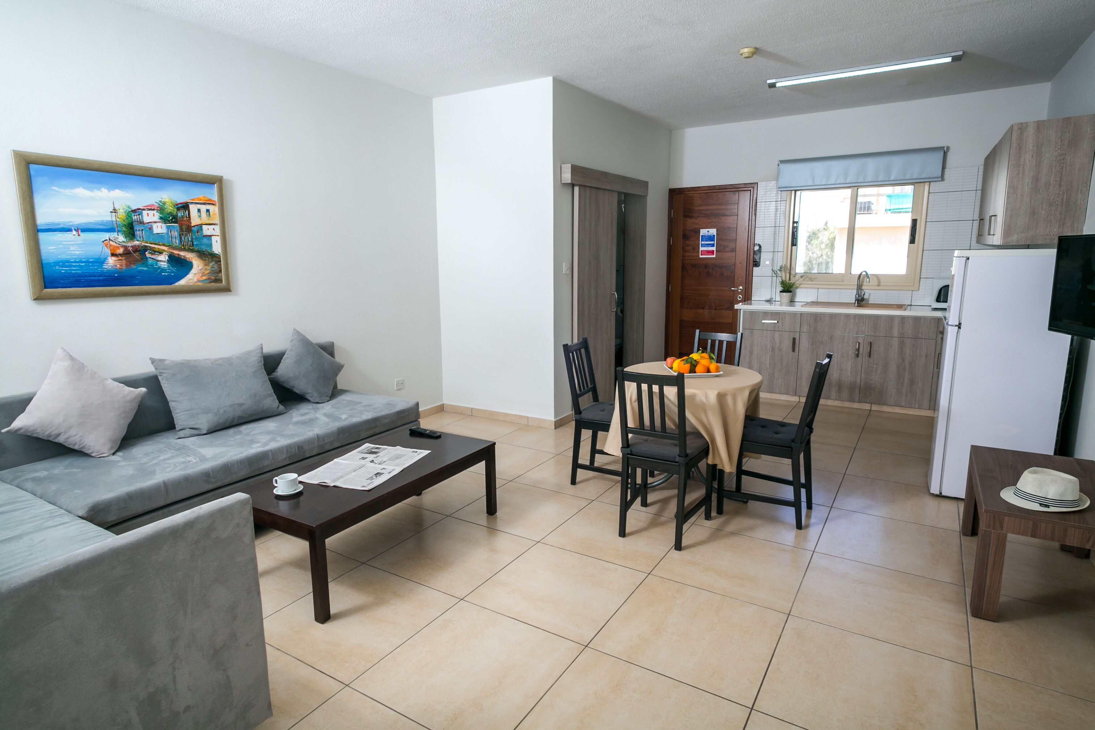 A modern living room with a sofa, coffee table, and a dining area with chairs and a table. There is a kitchen area with cabinets and a refrigerator.