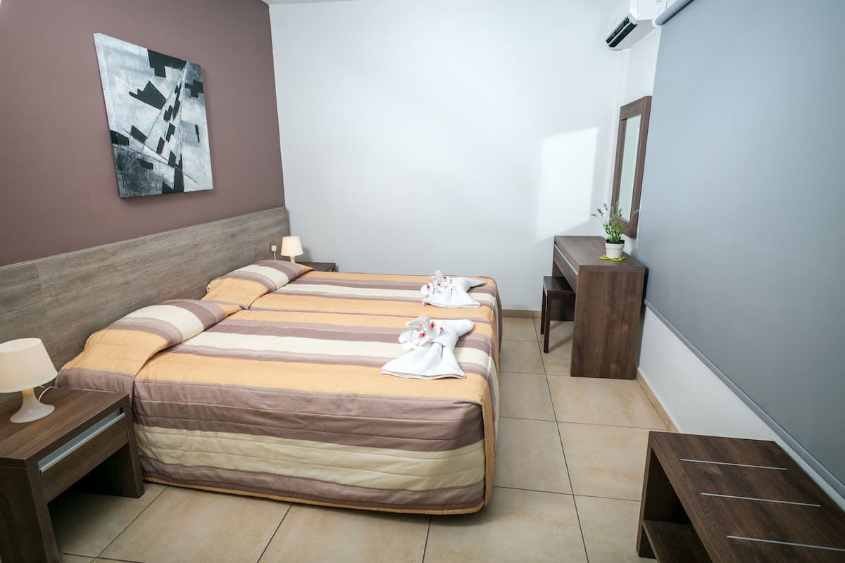 Una habitación de hotel con una cama grande, una mesita de noche con lámpara, un escritorio pequeño con espejo y un aire acondicionado instalado en la pared.