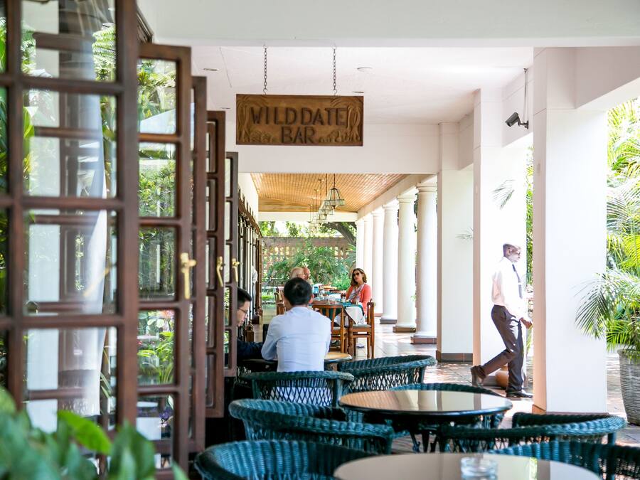 An outdoor seating area with wicker chairs and tables, a sign reading "Wilddate Bar", and a view of greenery.