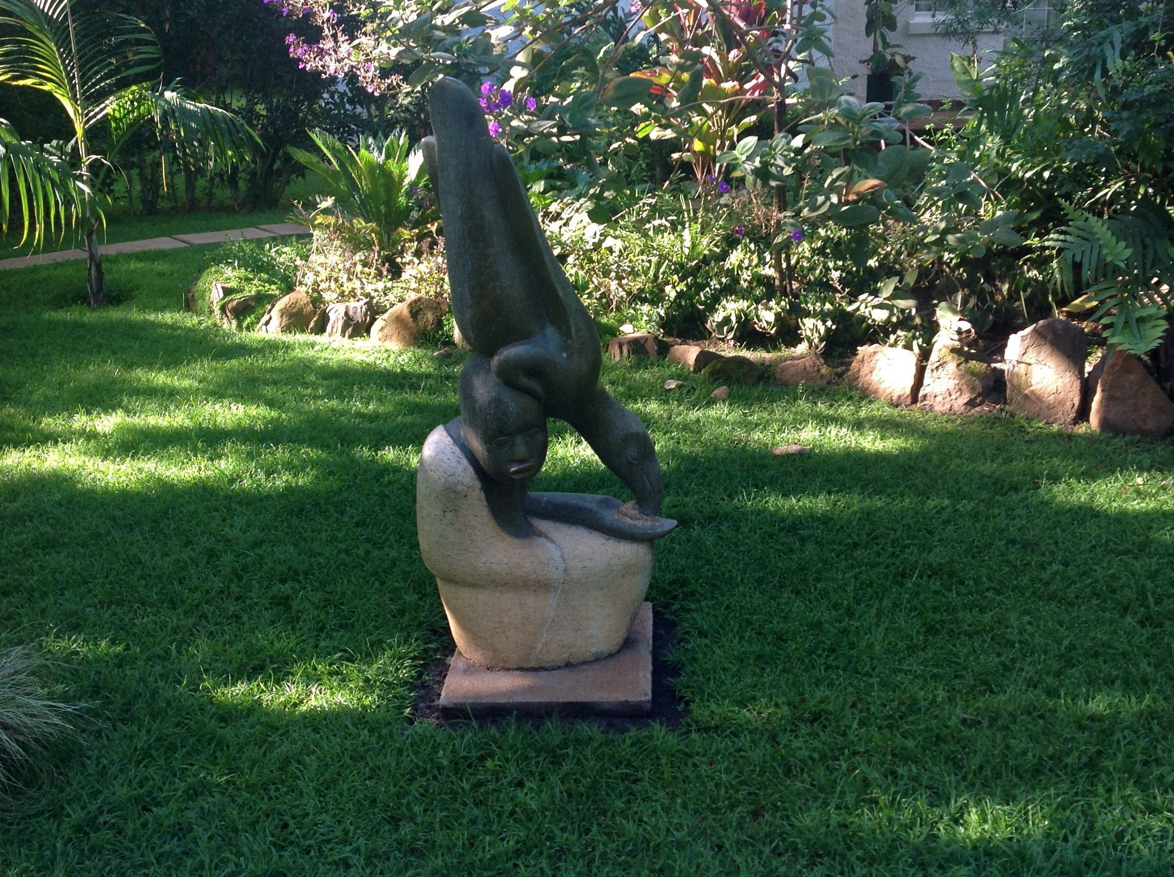A sculpture of a person in a dynamic pose on a pedestal in a garden with grass, rocks, and various plants.