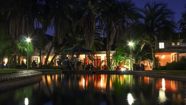 A nighttime view of a hotel with a pool reflecting lights, surrounded by palm trees and a well-lit outdoor seating area.