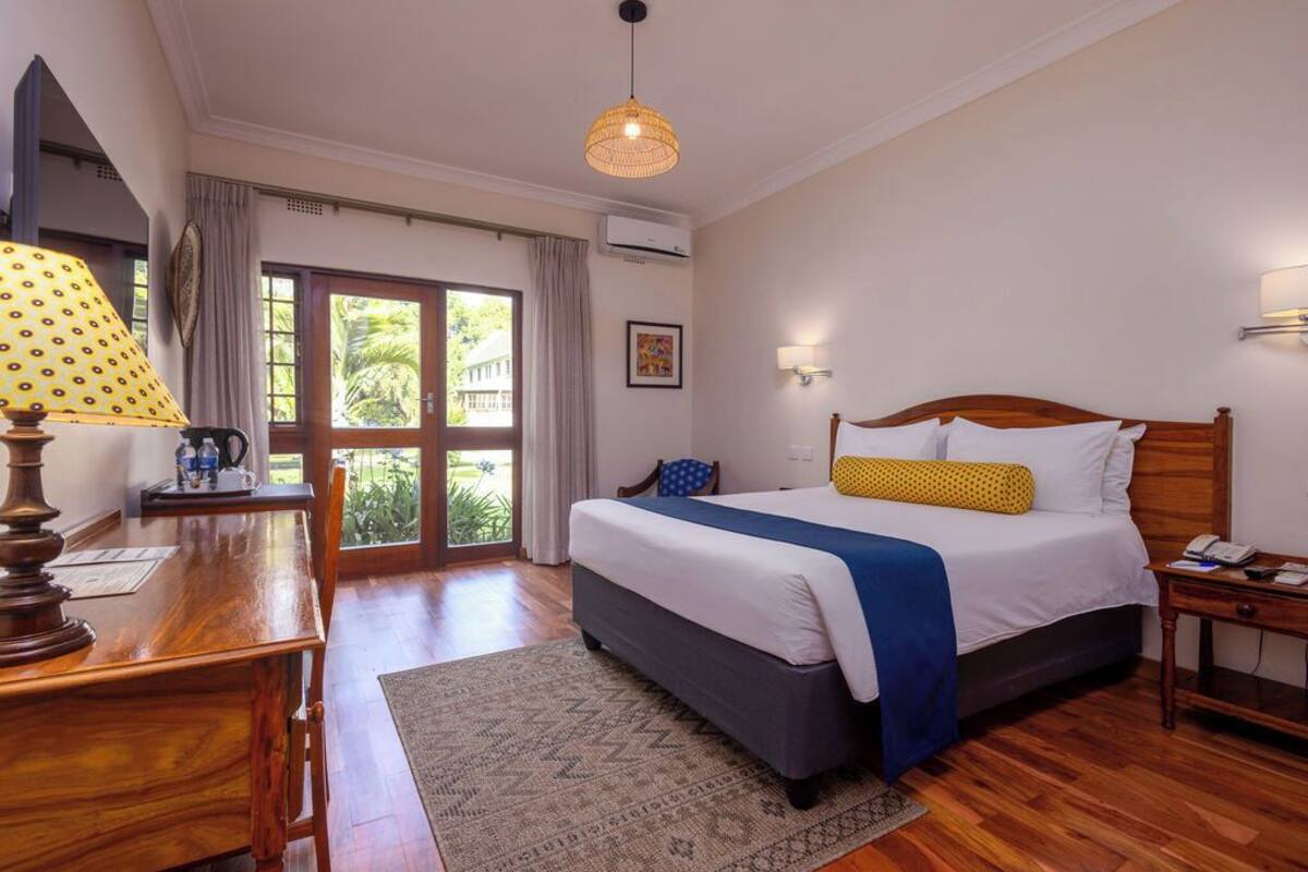 A bedroom with a large bed, a wooden desk, a lamp, a nightstand, and a view of greenery outside.