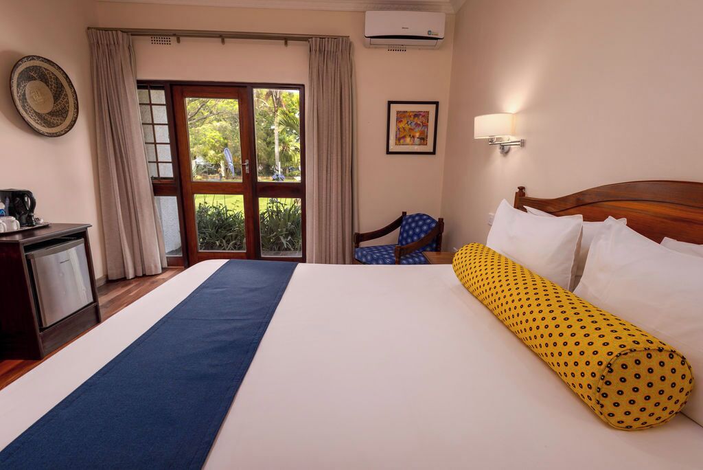 A hotel room with a large bed, a desk with a coffee maker, a chair, a window with curtains, and a wall-mounted air conditioner.