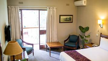 A hotel room with a bed, a desk, two chairs, a table, a lamp, a plant, a painting, and a sliding glass door.