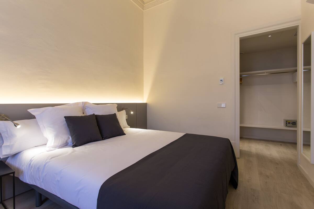 Een net opgemaakt bed met wit en donker beddengoed, een nachtkastje en een inloopkast met planken.