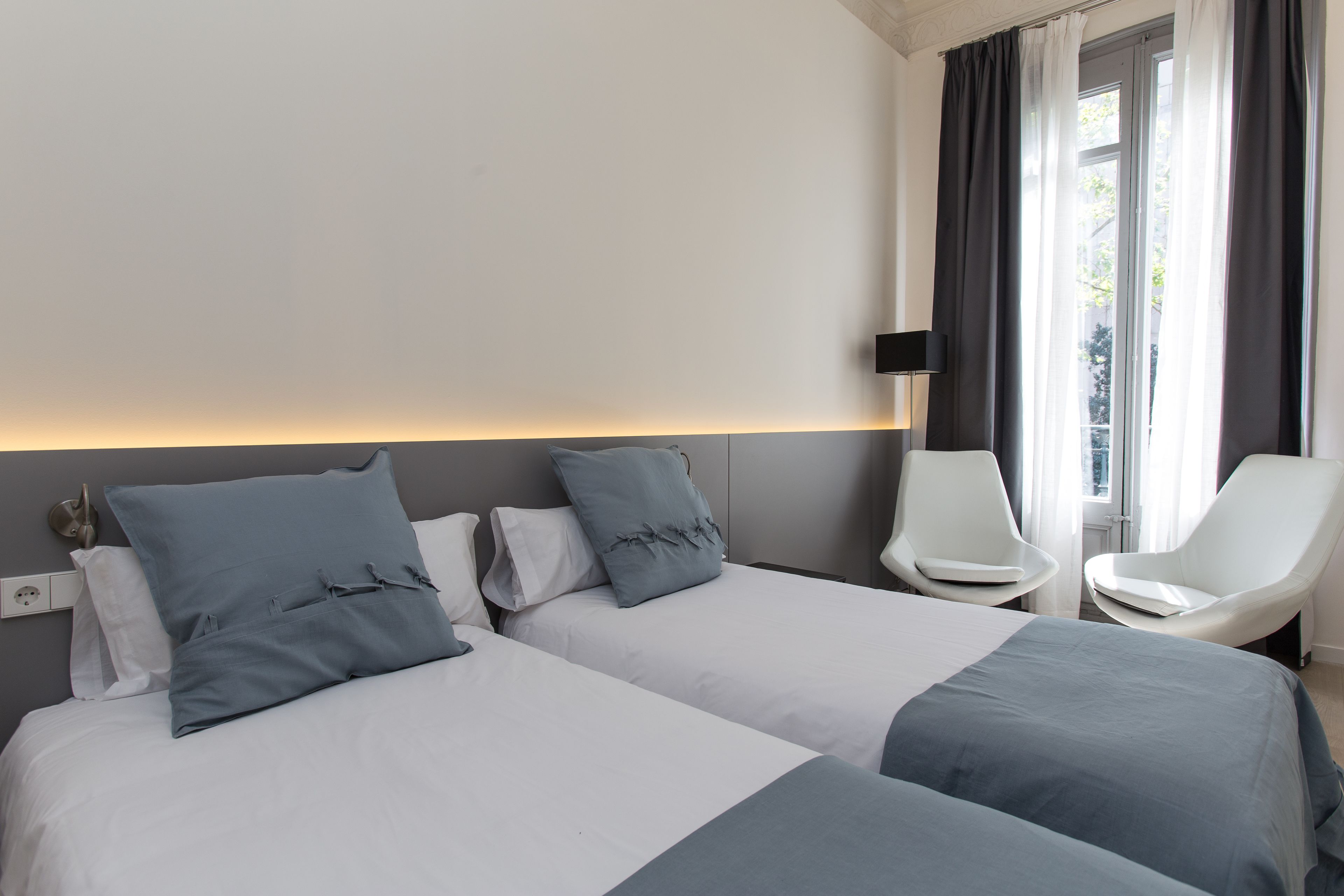 Habitación de hotel con dos camas, una zona de estar con sillas, una lámpara de pared y una ventana con cortinas.
