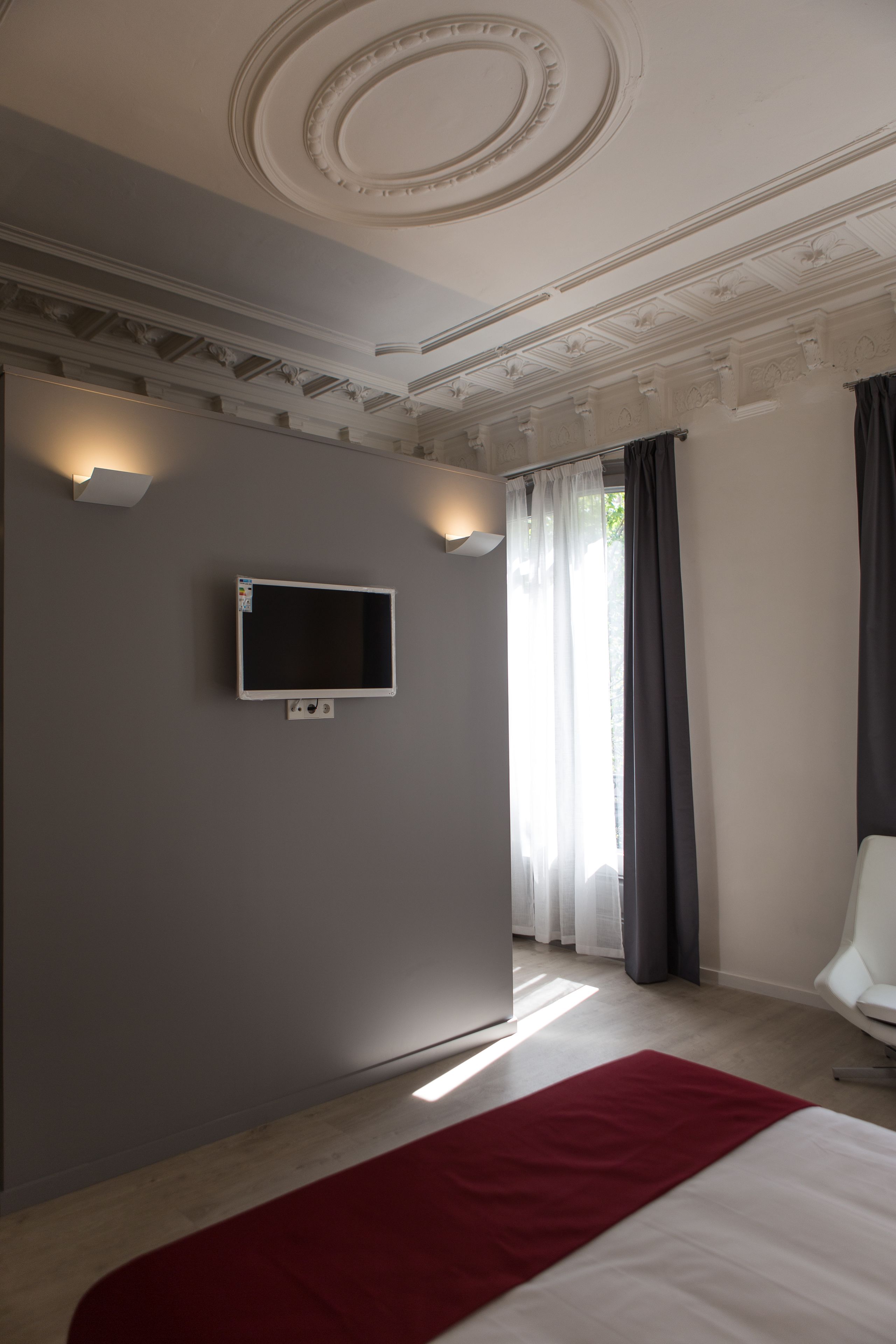 A modern hotel room with a flat-screen TV mounted on the wall, a large bed with a red throw, and a white chair.