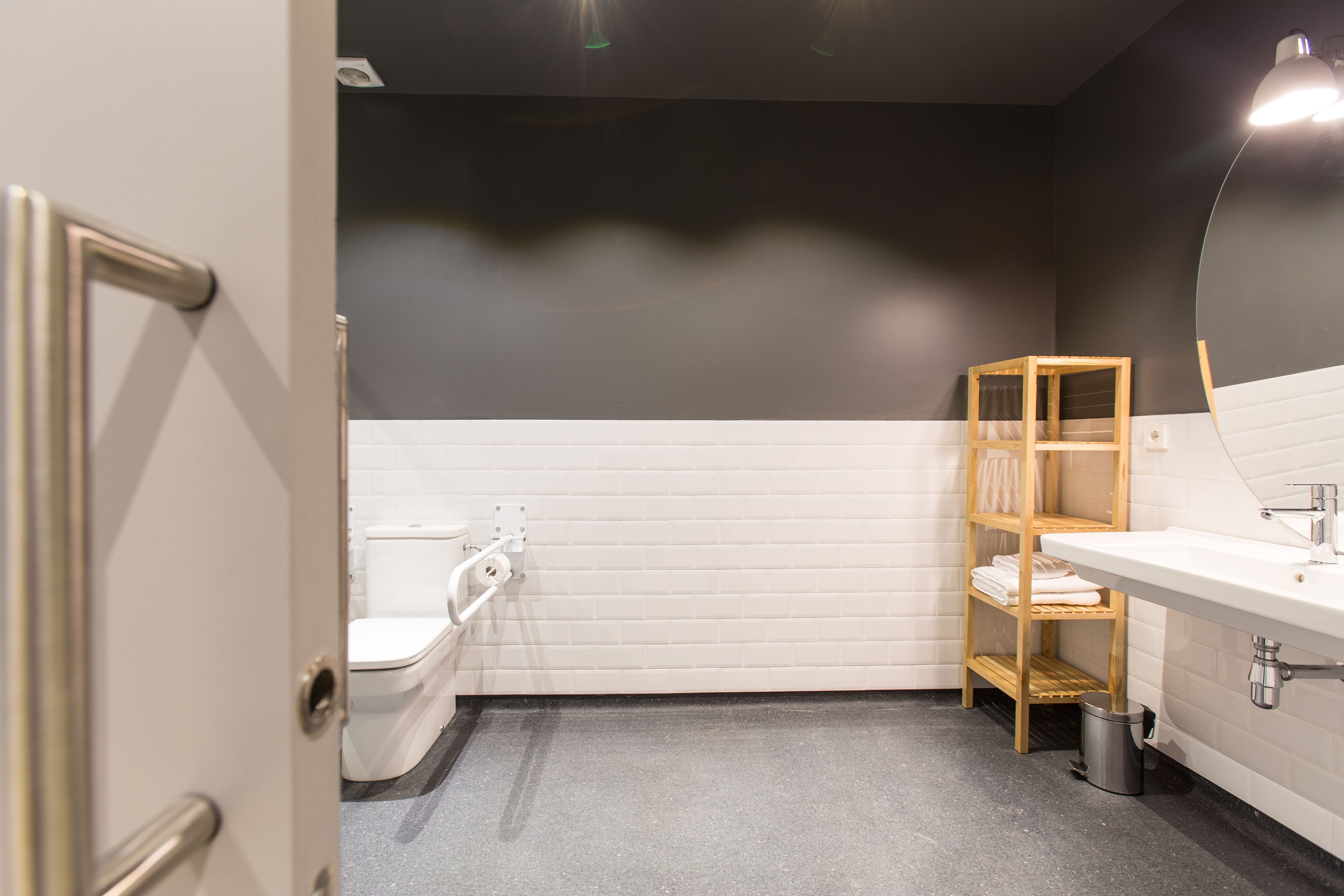 A bathroom with a white toilet, a sink with a mirror, a shelf with towels, and a trash can.