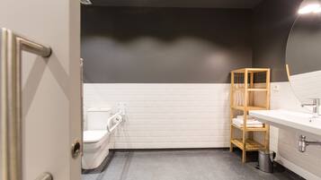 A bathroom with a white toilet, a sink with a mirror, a shelf with towels, and a trash can.