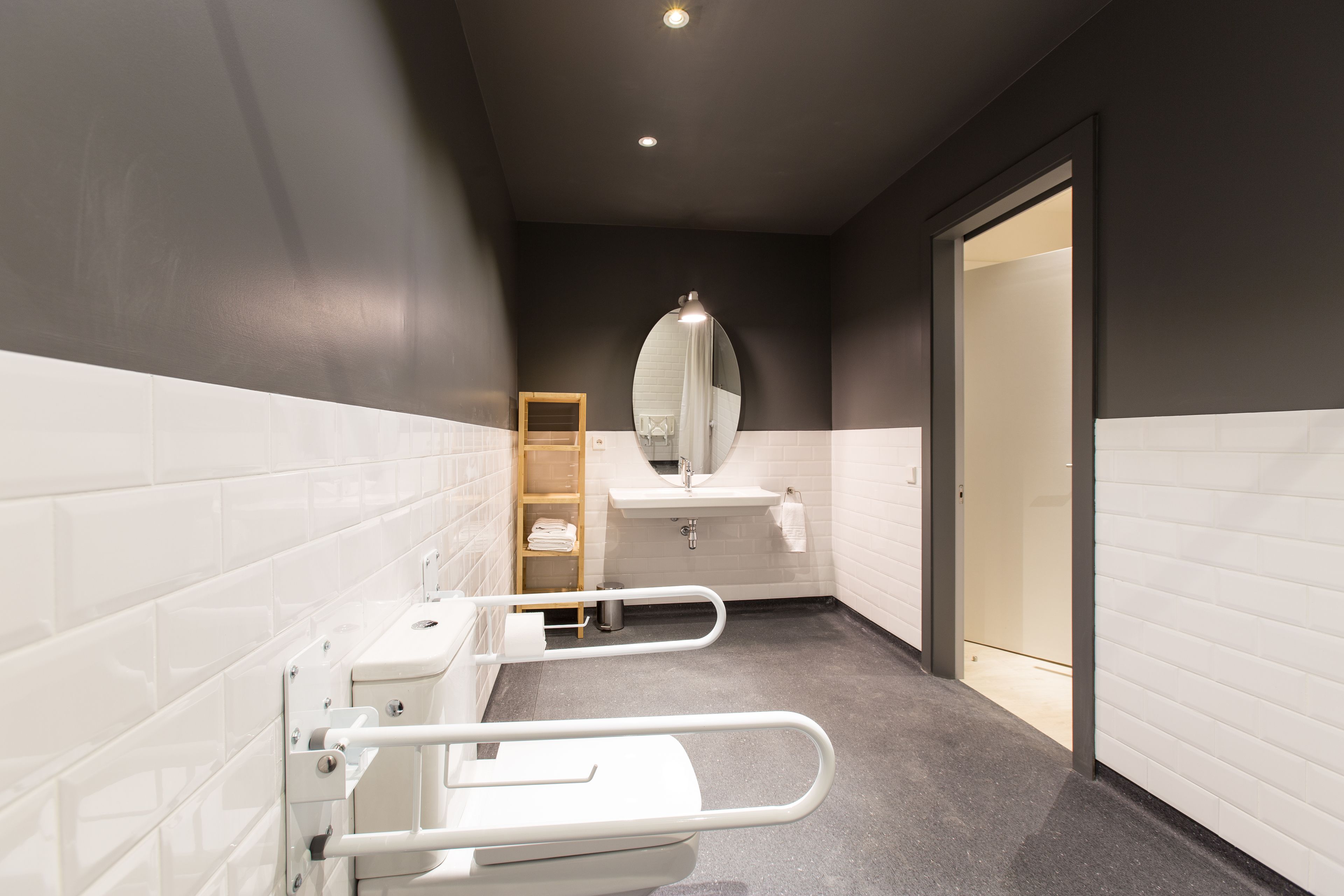 Un baño moderno con paredes de azulejos blancos, suelo gris, inodoro blanco con barra de apoyo, lavabo con espejo y una puerta que da a otra habitación.