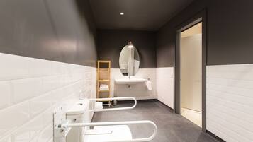 Un baño moderno con paredes de azulejos blancos, suelo gris, inodoro blanco con barra de apoyo, lavabo con espejo y una puerta que da a otra habitación.