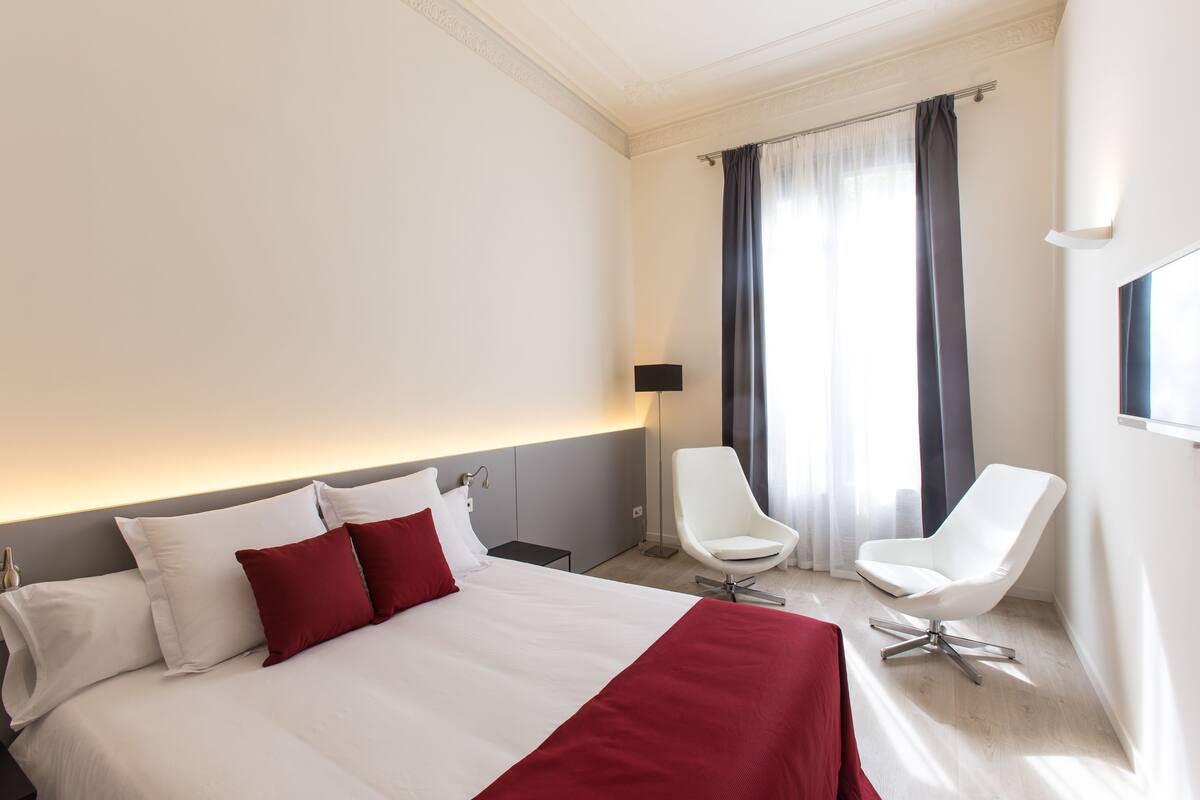 Een hotelkamer met een groot bed, een witte fauteuil, een nachtkastje en een raam met gordijnen.