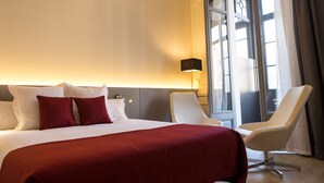 Una habitación de hotel con una cama grande, dos sillas blancas, una mesita de noche y una ventana con cortinas.
