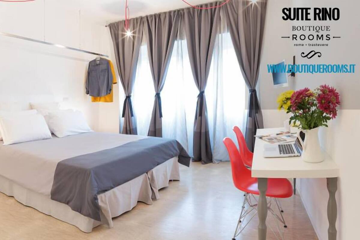 A modern hotel room with a bed, a desk with a laptop, a red chair, and a window with curtains.