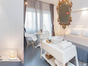 Una camera d'albergo moderna con un letto, un comodino con uno specchio, un piccolo tavolo rotondo con sedie e una finestra con tende.