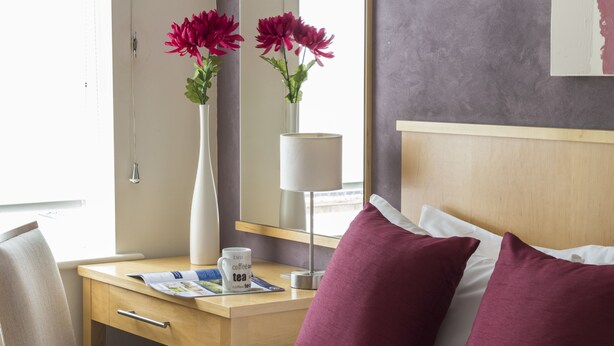 A hotel room with a bed, a desk, a chair, a vase with flowers, a lamp, and a magazine on the desk.