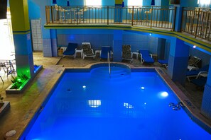 An indoor swimming pool with blue tiles, lounge chairs, and a balcony with railings.