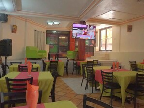 A restaurant with multiple tables covered in yellow and red tablecloths, green chairs, and a television screen displaying a cooking show.