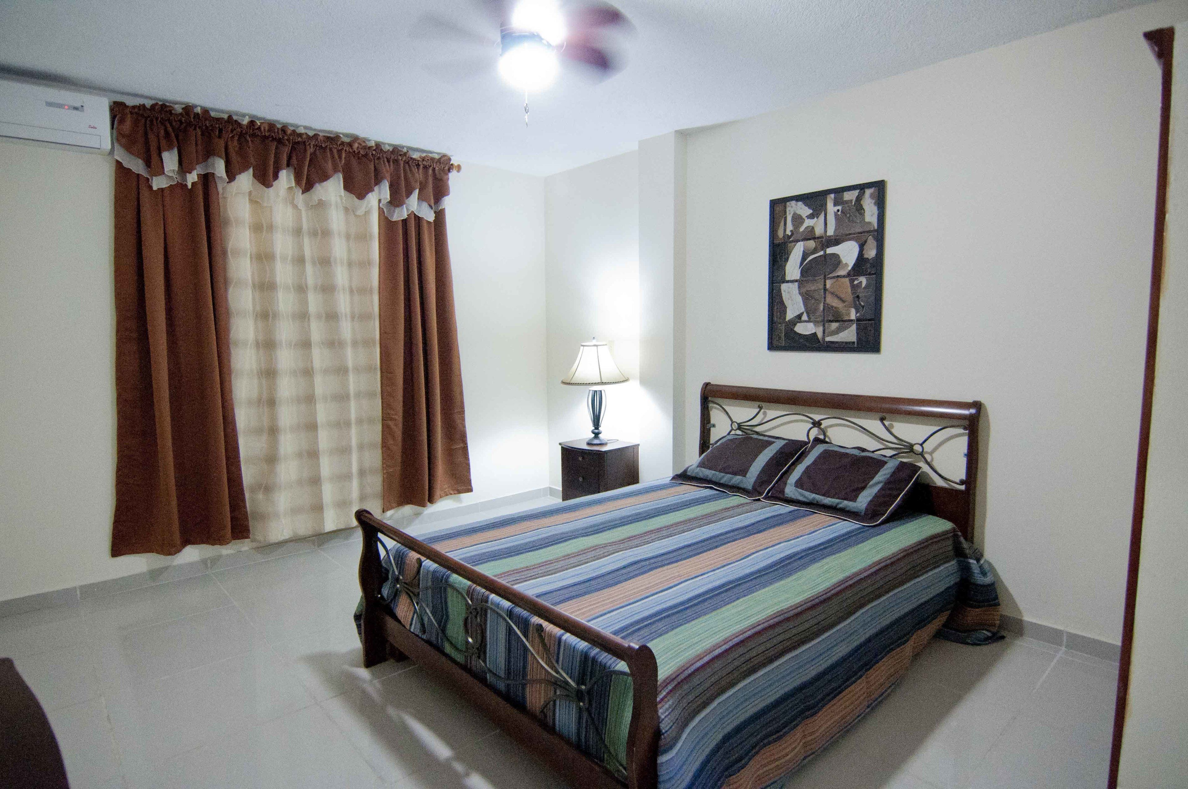 Un dormitorio con cama, cortinas, una mesita de noche, una lámpara y una obra de arte en la pared.