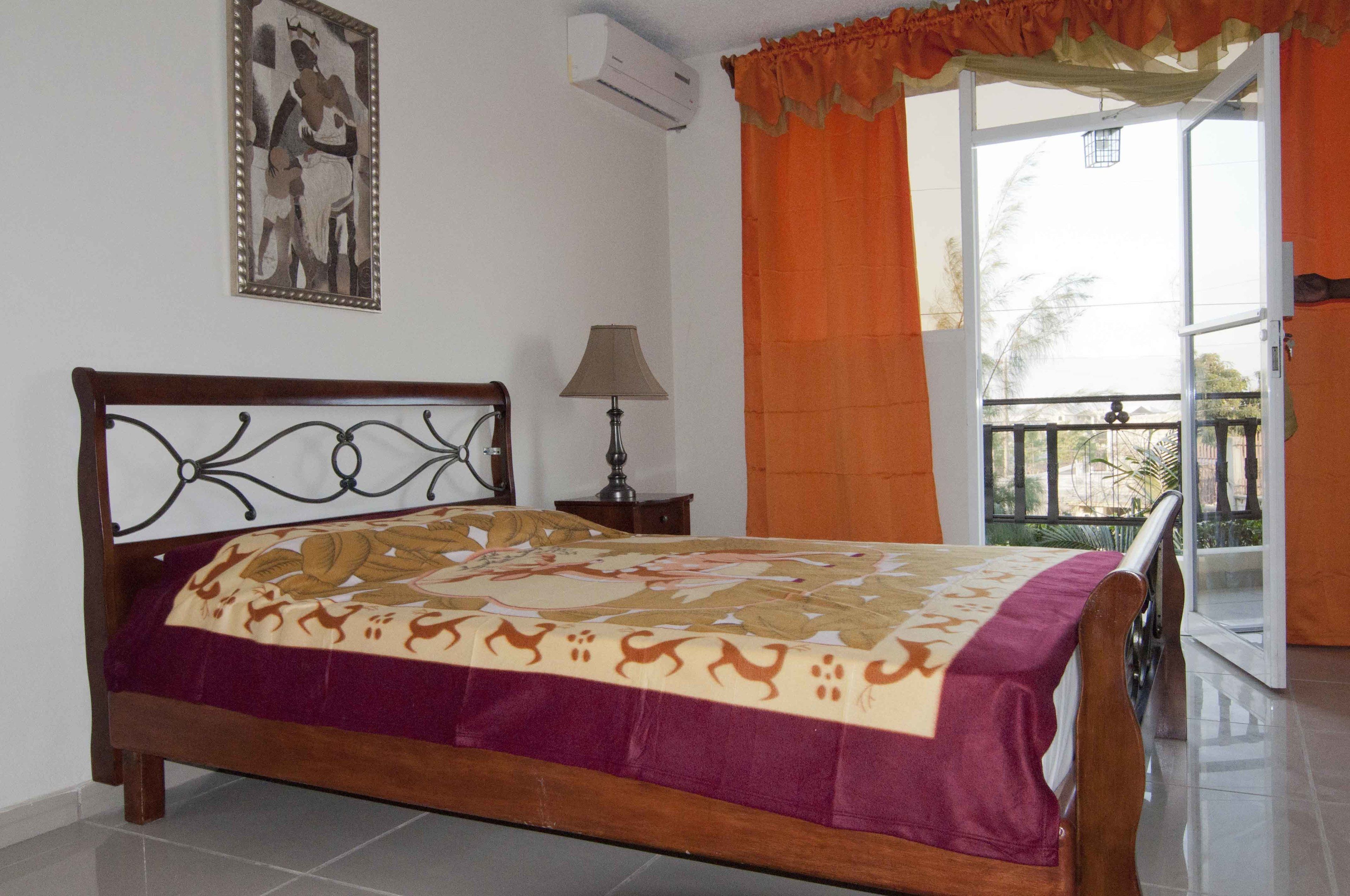 Un dormitorio con una cama de madera, una mesita de noche, una lámpara, un cuadro en la pared y un balcón con cortinas naranjas.