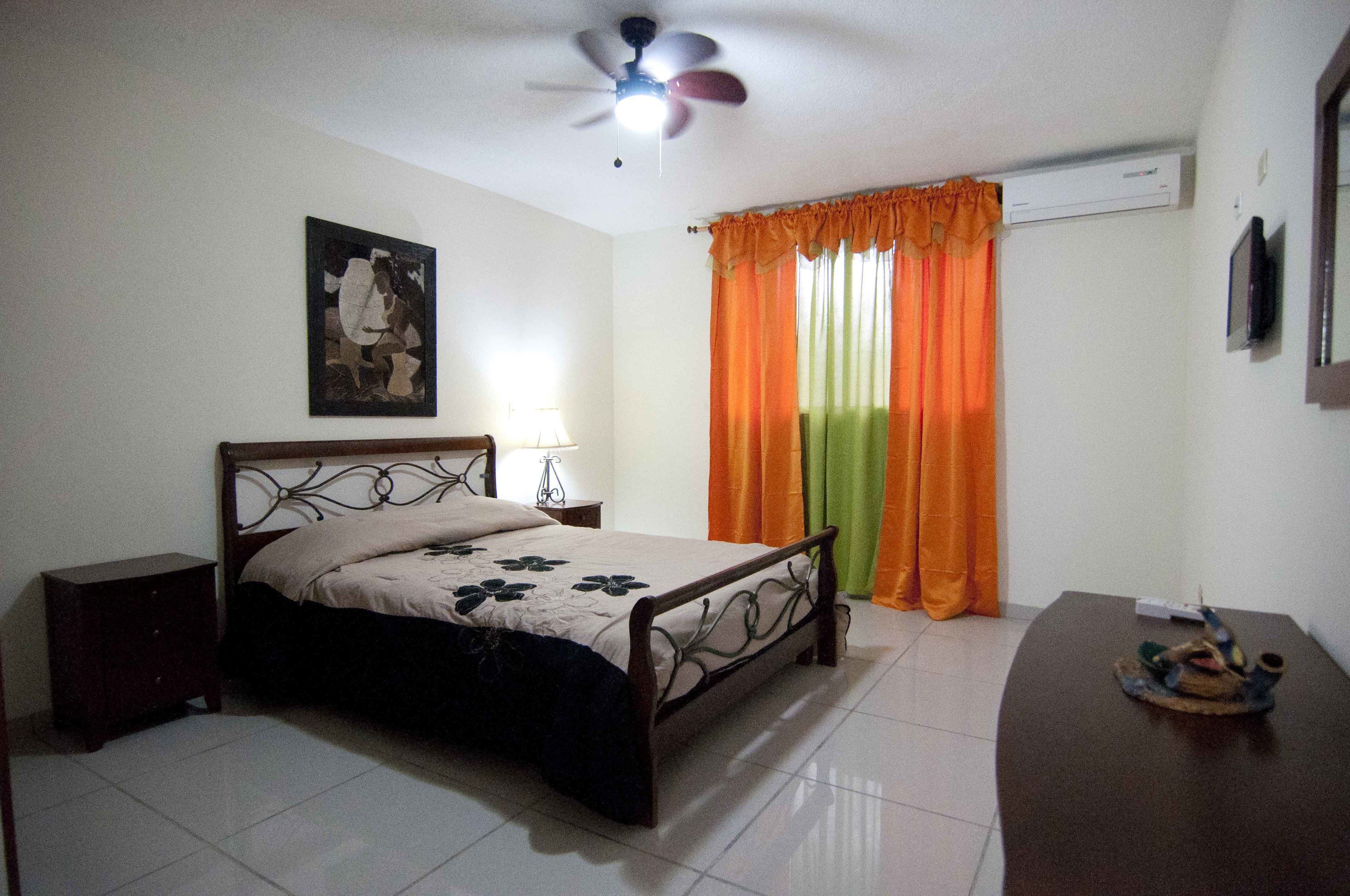 A bedroom with a bed, a nightstand, a ceiling fan, an air conditioner, and a window with orange curtains.