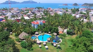 An aerial view of a resort with multiple swimming pools surrounded by lush greenery and palm trees, set against a backdrop of a coastal town with buildings and mountains. - Hotel & Temazcal Hacienda de Melaque (San Patricio Melaque)