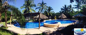 Una piscina con una estructura de techo de paja, rodeada de palmeras y sillas de descanso.