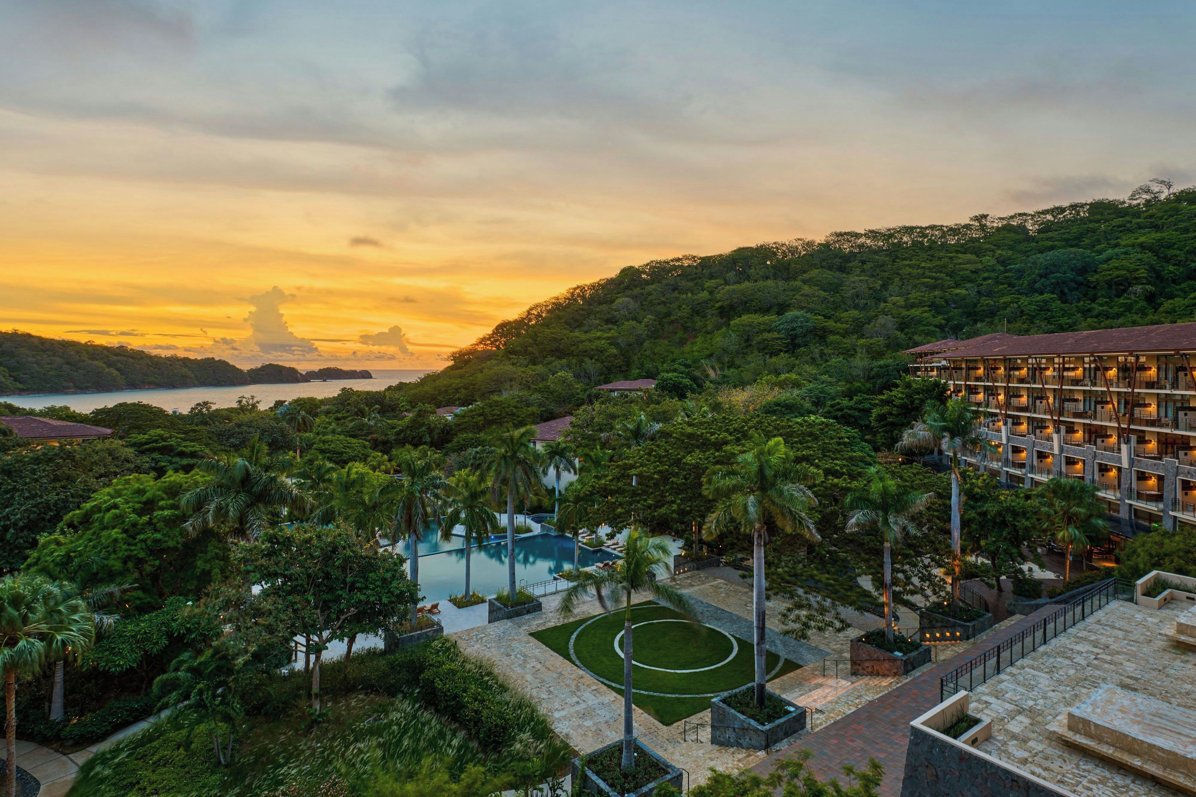 Un resort con piscina, palmeras y vista al mar al atardecer.