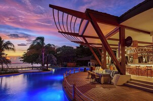 A resort pool area with a wooden deck, a bar, and a view of the ocean at sunset.