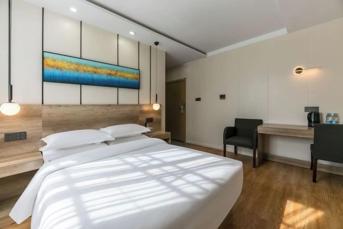 A modern hotel room with a large bed, a desk, and a chair. There is a painting on the wall and a ceiling light.