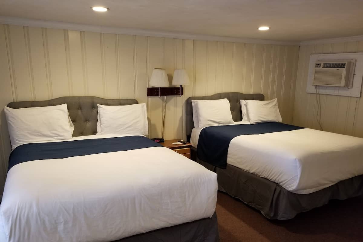 Two single beds with grey headboards and white bedding, a nightstand with a clock, and a wall-mounted air conditioner.