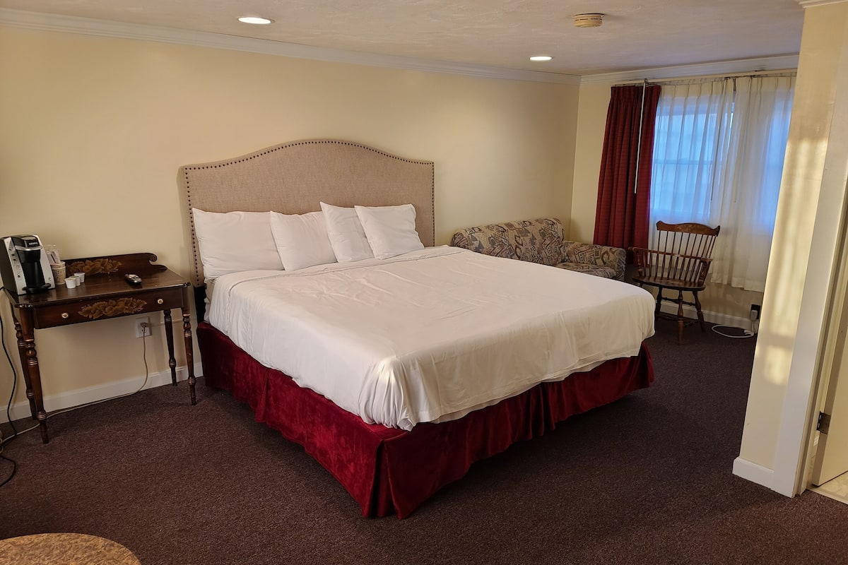 A neatly made bed with a red skirt, a wooden nightstand with a coffee maker, a patterned armchair, and a wooden chair.