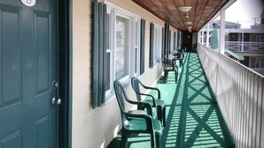 Un couloir abrité, avec des chaises et des tables vertes, des poutres en bois au plafond et une vue sur les bâtiments en arrière-plan.