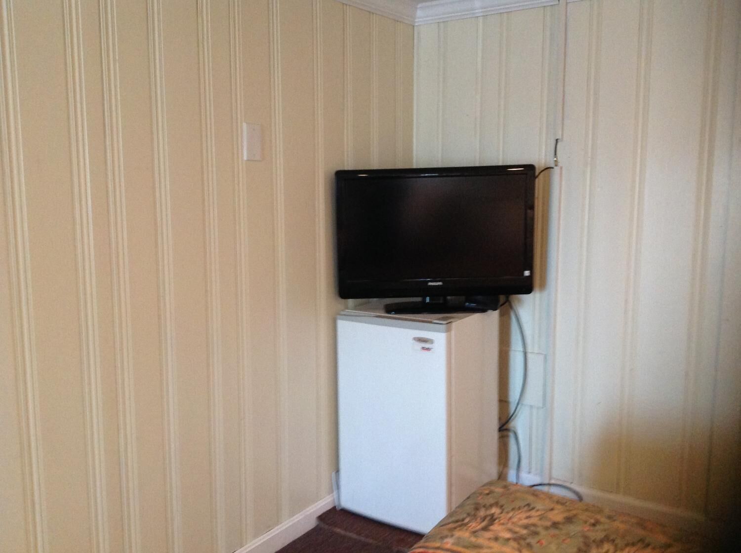 A room with a television mounted on a wall, a small white refrigerator, and a patterned bench.