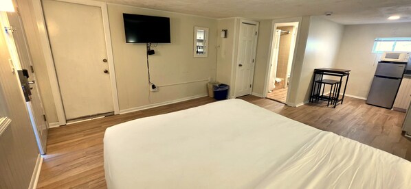 A room with a bed, a TV mounted on the wall, a small kitchen area with a refrigerator, and a door leading to another room. - The Tides Motel (Hampton)
